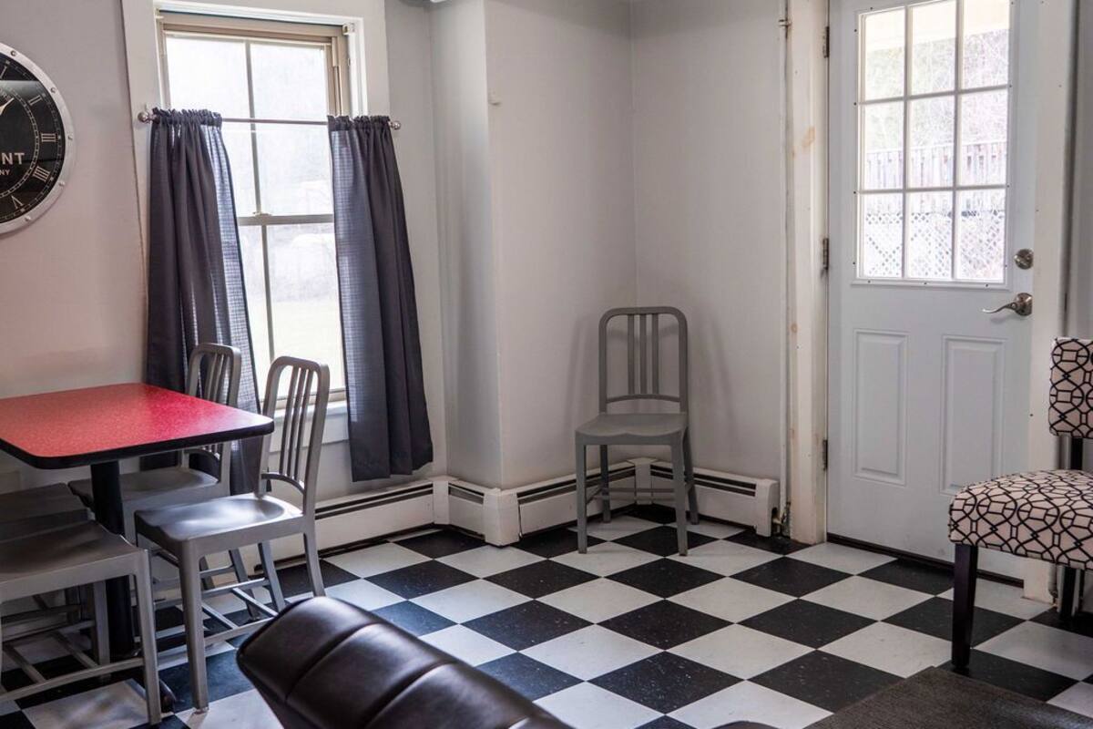 Una habitación con piso a cuadros, una mesa roja, dos sillas grises, un banco de cuero negro, otra silla gris, una puerta blanca y una ventana con cortinas.