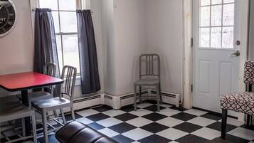 A room with a checkered floor, a red table, two grey chairs, a black leather bench, a grey chair, a white door, and a window with curtains.