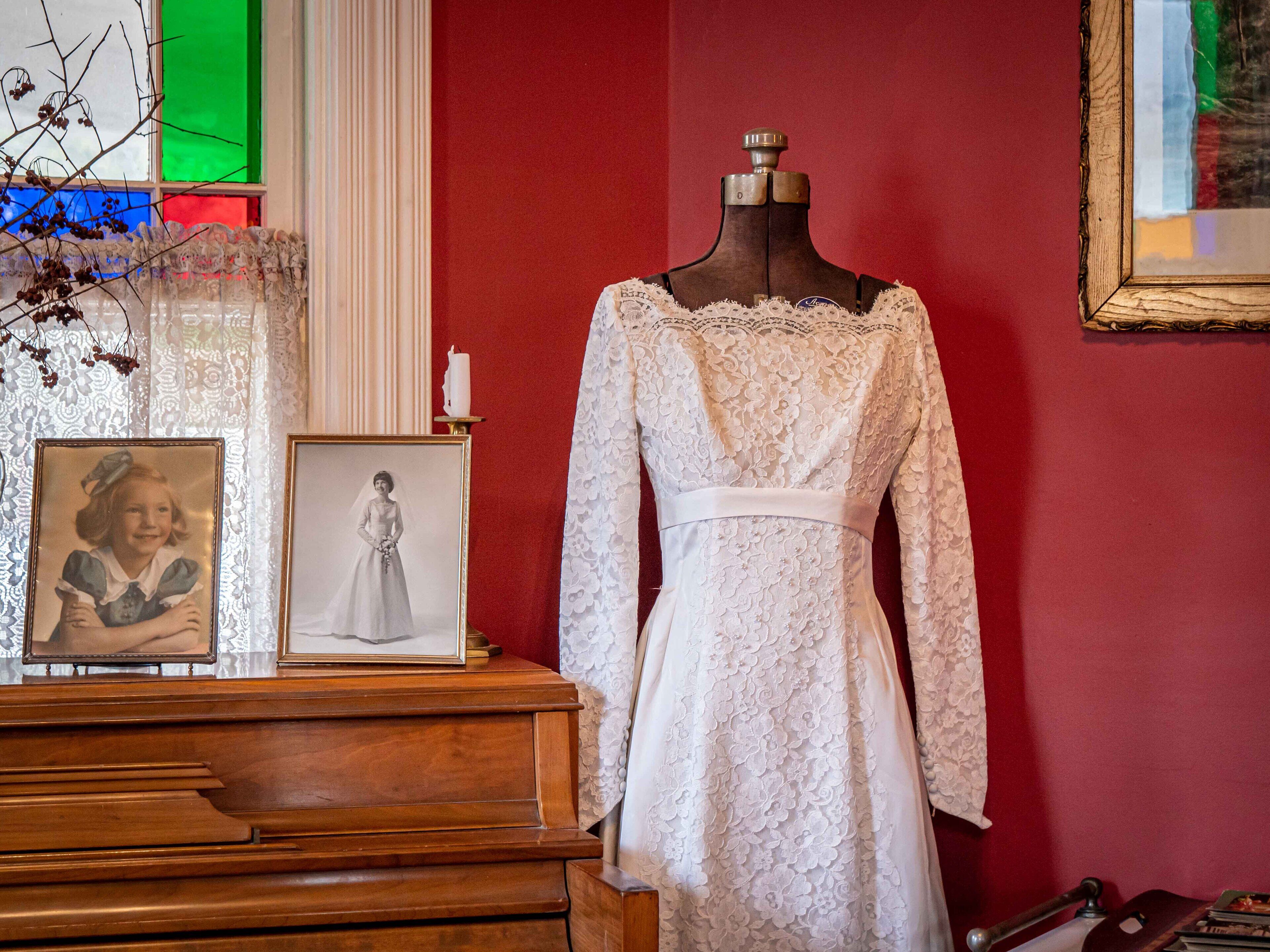 Un maniquí vestido con un traje de novia blanco de encaje, con falda larga y corpiño entallado, exhibido sobre un tocador de madera frente a una pared roja.