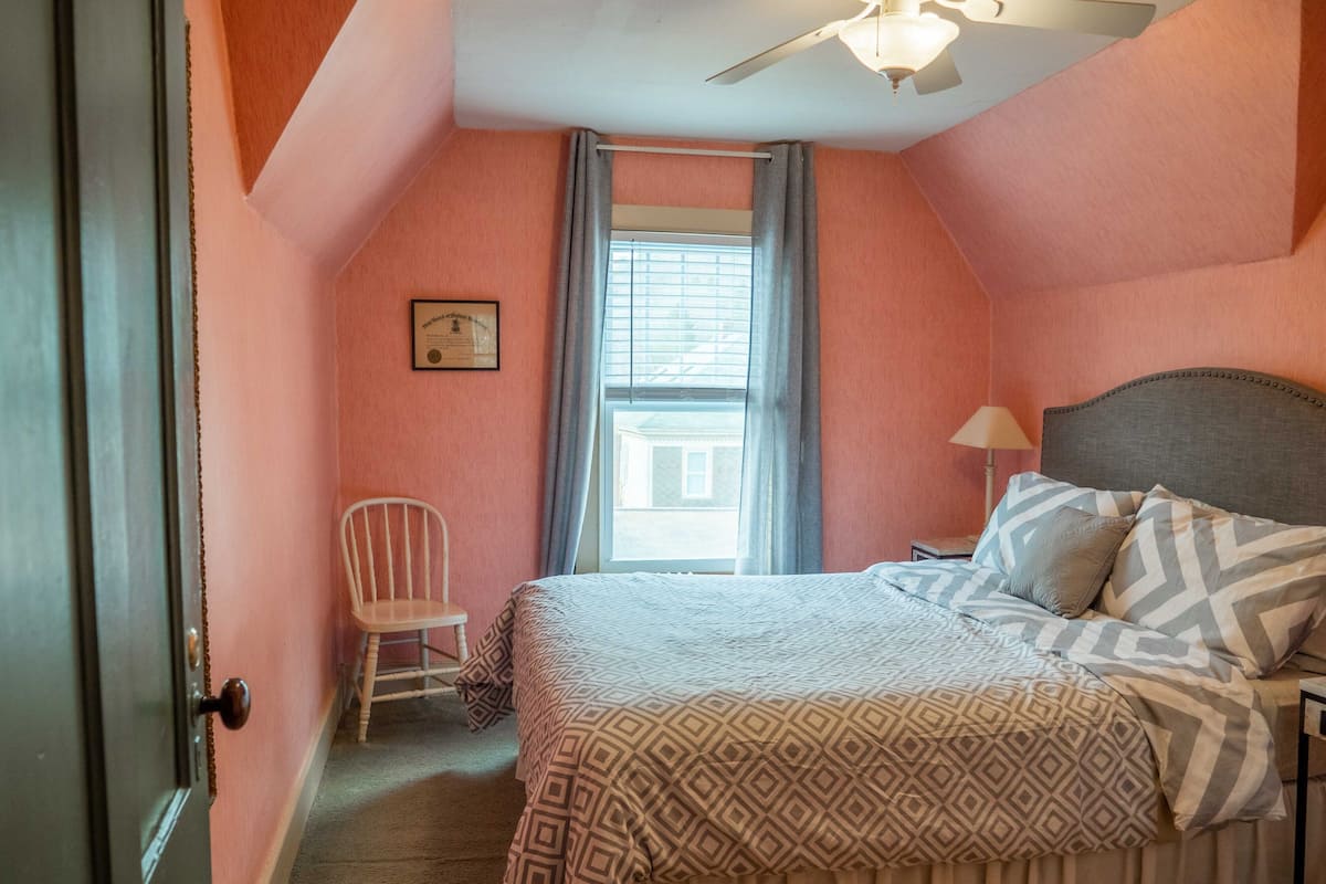 Un dormitorio con cama, una silla, un ventanal con cortinas y un ventilador de techo.