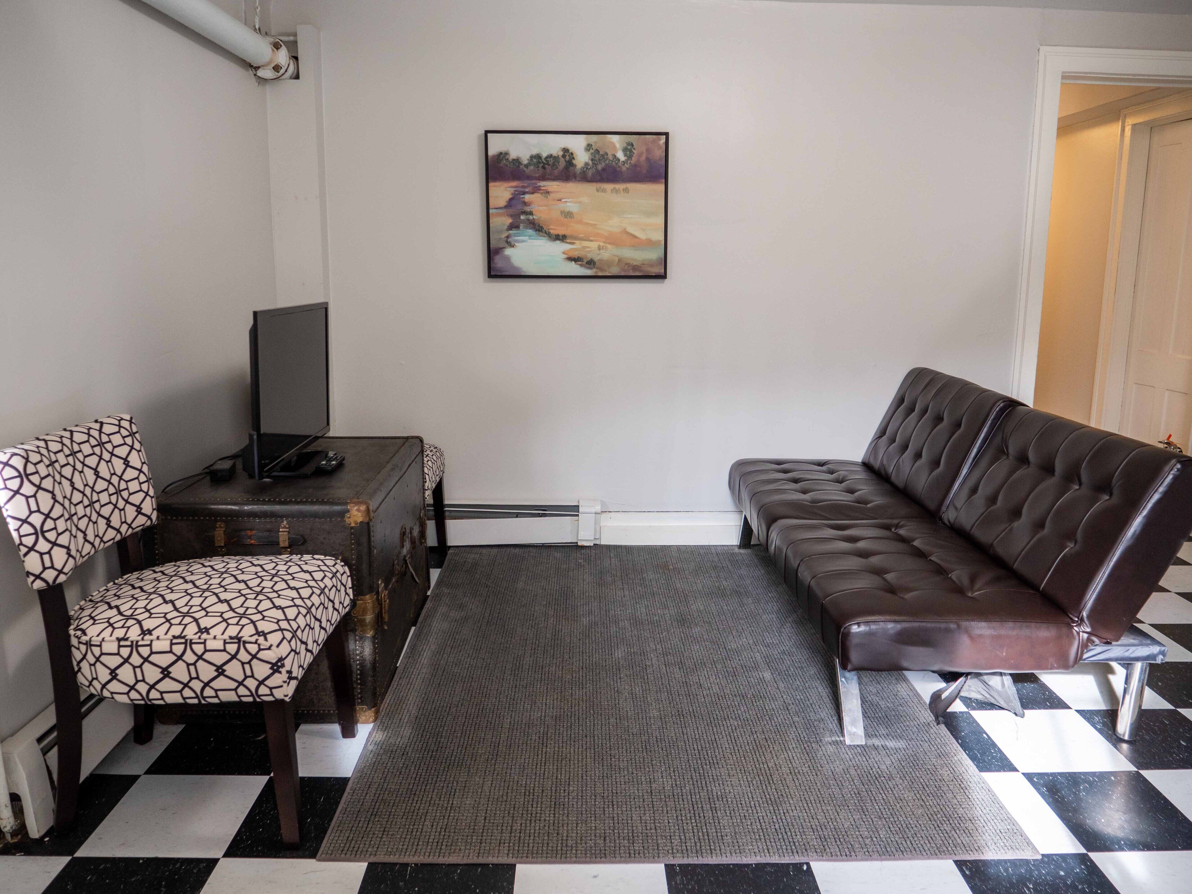 A living room with a checkered floor, a brown leather sofa, a patterned armchair, a small table with a TV, and a painting on the wall.