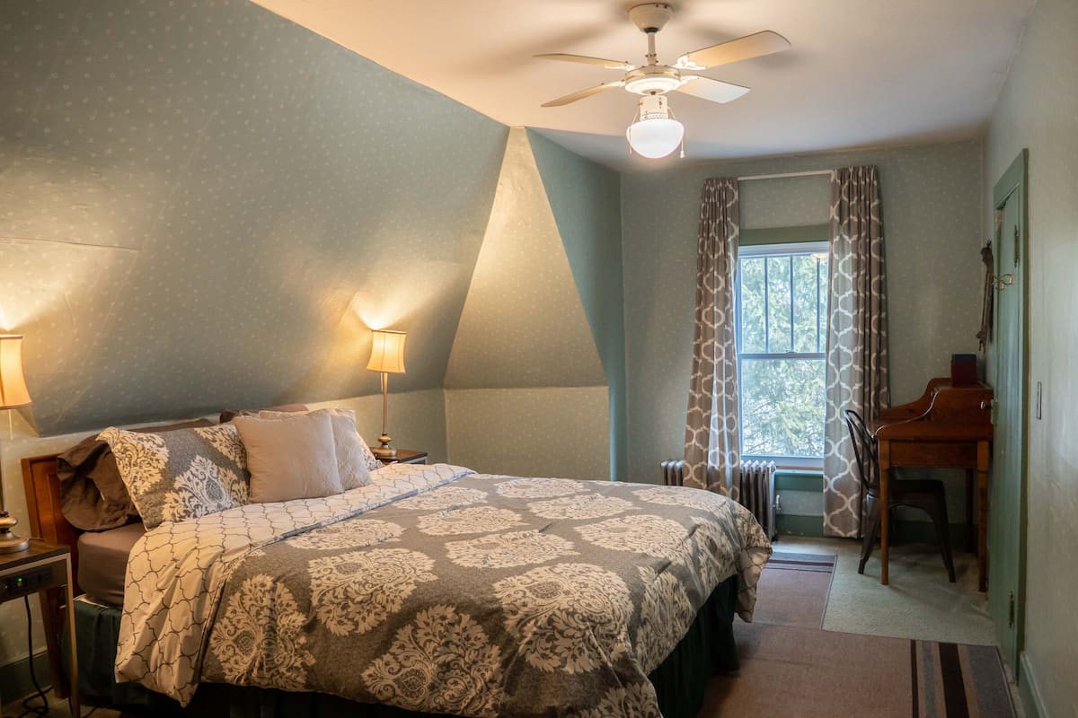 Un dormitorio con cama, mesitas de noche, escritorio, ventilador de techo y ventana con cortinas.