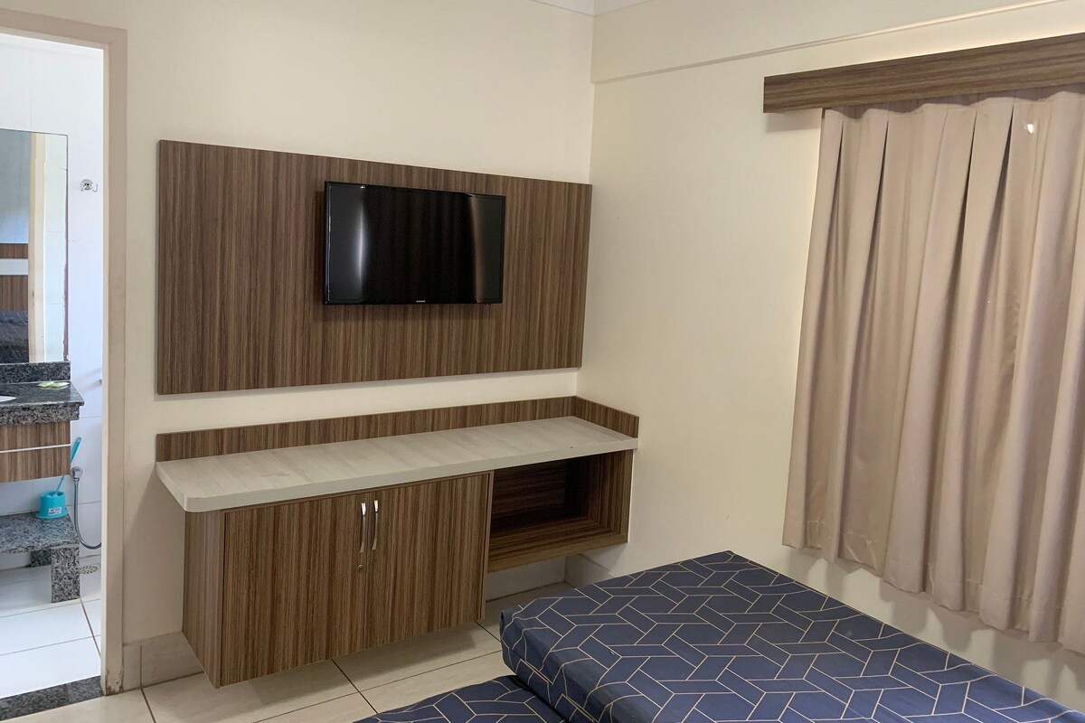 Un dormitorio con una televisión montada en una pared de paneles de madera, una cama con colcha estampada y una ventana con cortinas.