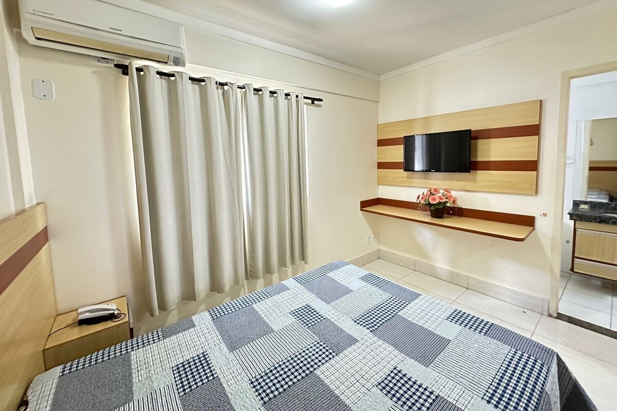 A bedroom with a bed, curtains, a TV mounted on a wooden shelf, a small table with a vase, and a kitchenette area.