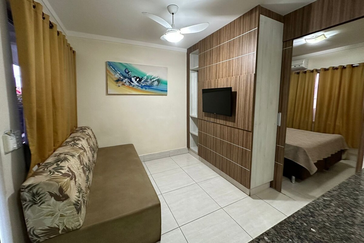 Una habitación con sofá, televisor montado en una pared de paneles de madera, ventilador de techo y cama con cortinas.