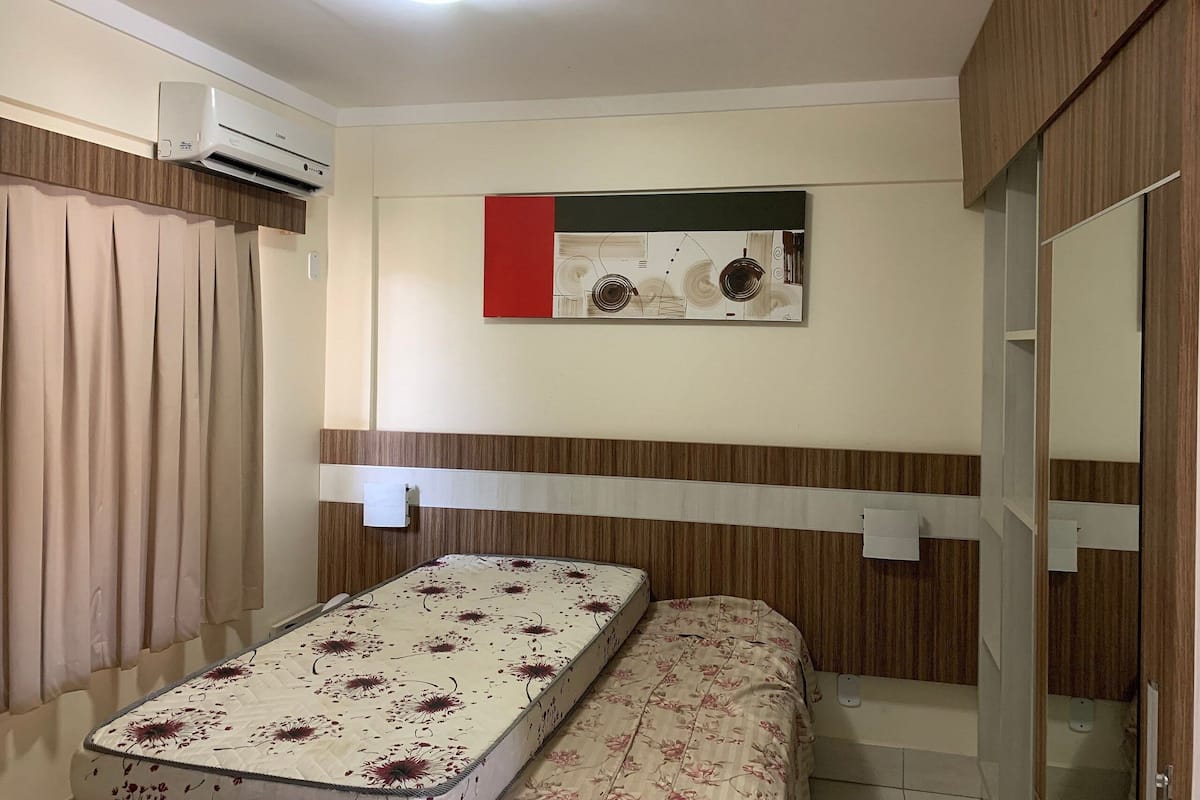 Quarto compacto com cama, janela pequena, ar-condicionado fixado na parede e espelho.