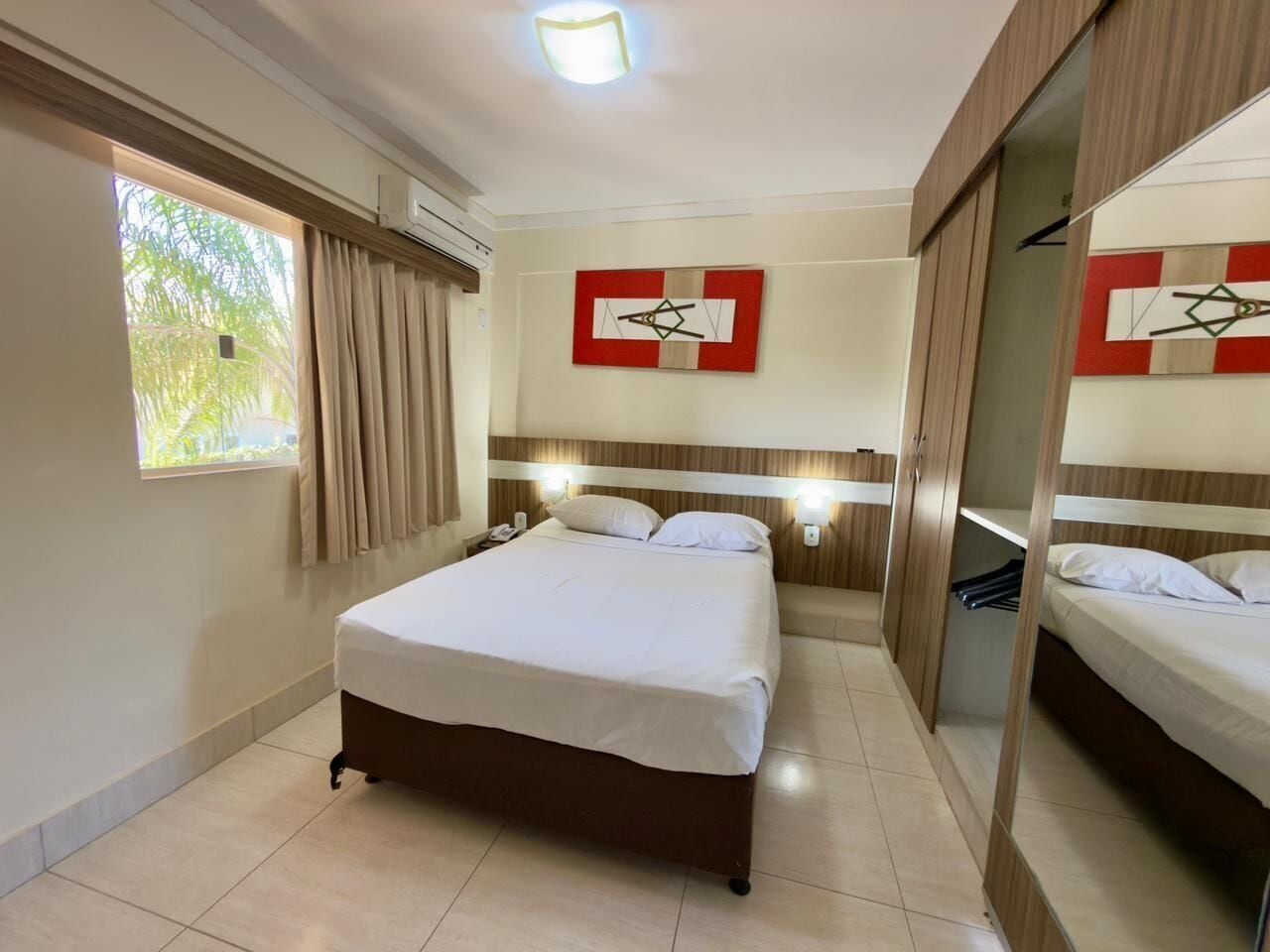 A hotel room with a large bed, a bedside table, a mirror, a window with curtains, and a wall-mounted artwork.