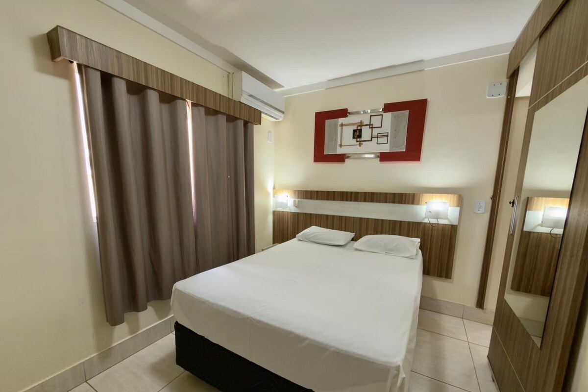 Una habitación de hotel con cama, cortinas, cabecero, espejo montado en la pared y mesita de noche.