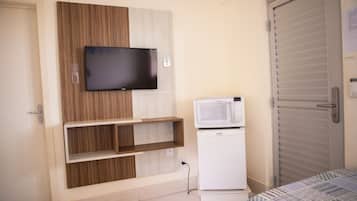 Quarto com televisão fixada numa parede revestida de madeira, micro-ondas sobre um armário branco e uma porta com puxador.