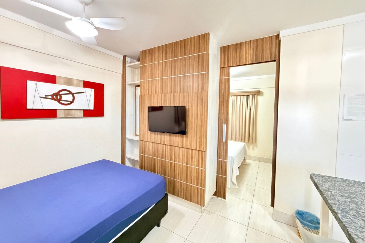 Un dormitorio con cama, televisor montado en una pared de paneles de madera, ventilador de techo y baño visible a través de una puerta.