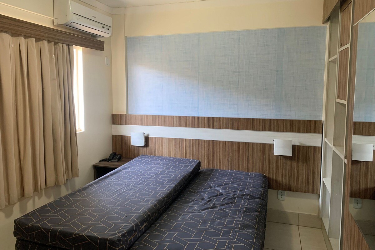 A small, compact room with a bed, a bedside table, a wall-mounted air conditioner, and a window with curtains.