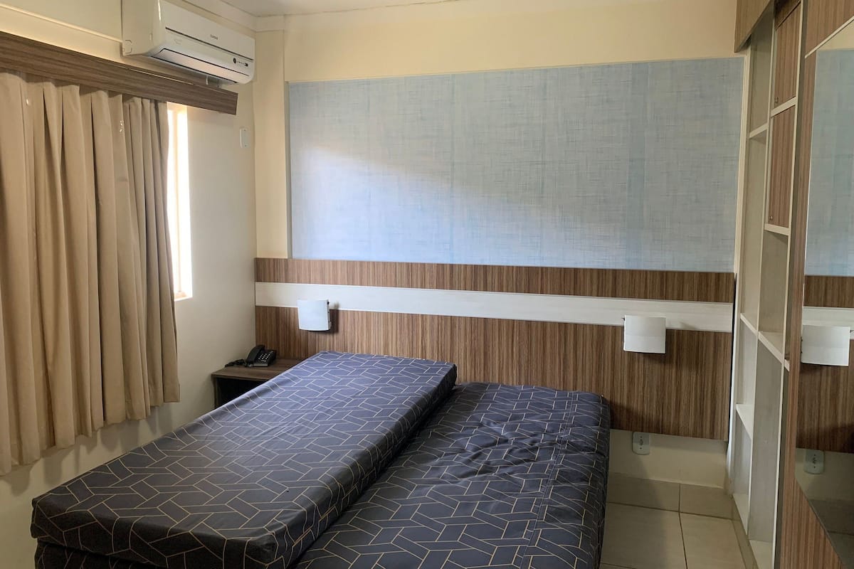 A small, compact room with a bed, a bedside table, a wall-mounted air conditioner, and a window with curtains.