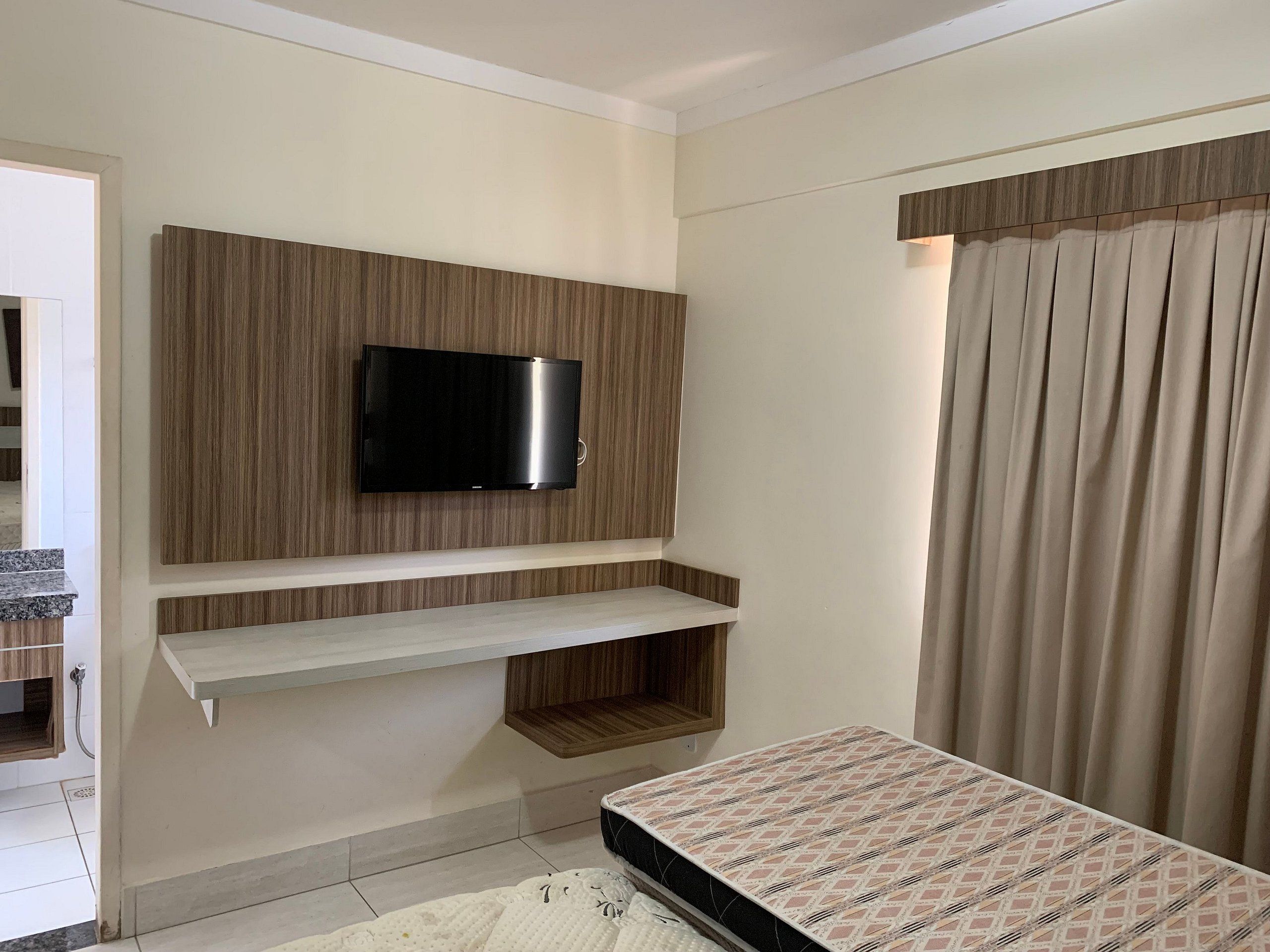 Quarto com televisão fixada em painel de madeira, cama com roupa de cama estampada e cortina na janela.