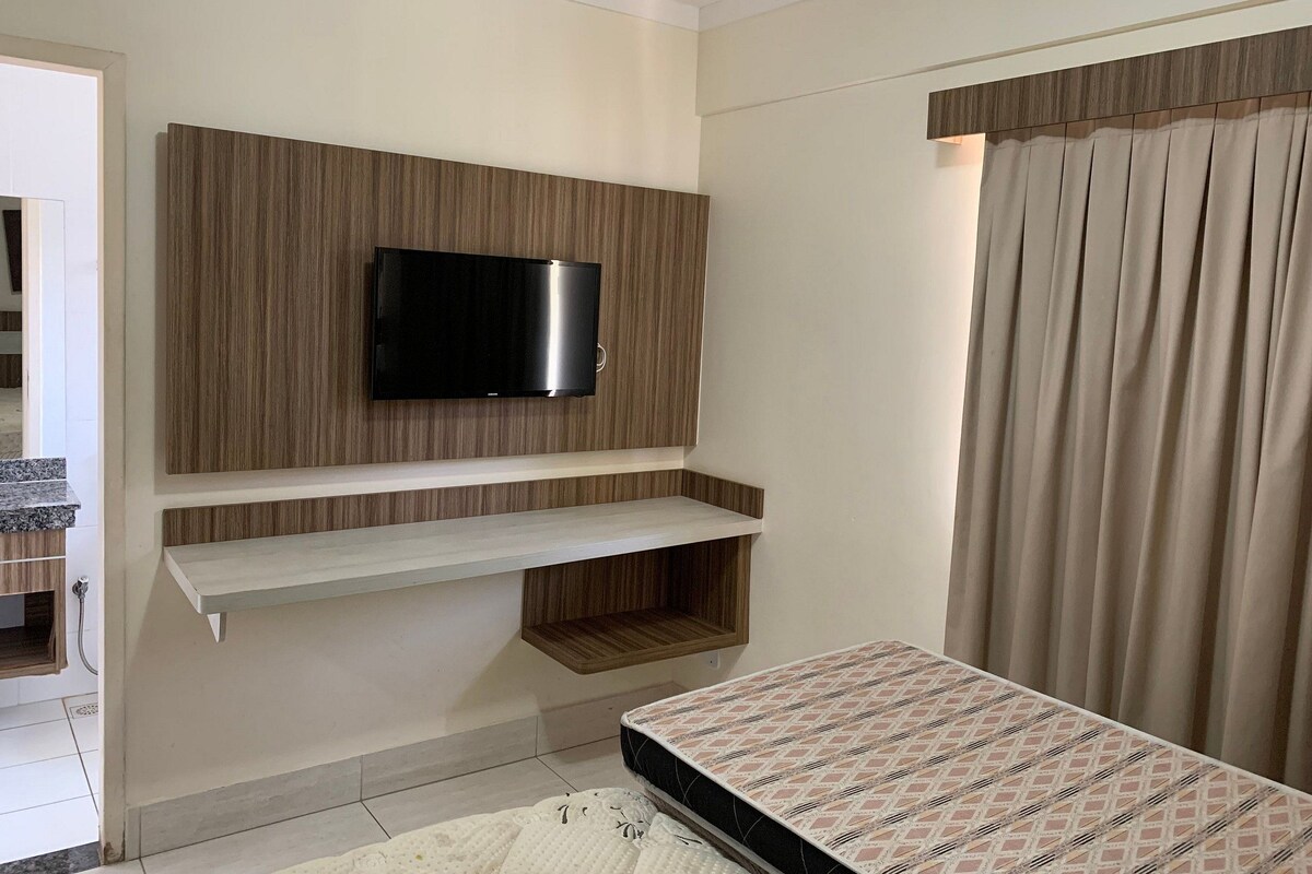 Un dormitorio con una televisión montada en una pared de paneles de madera, una cama con ropa de cama estampada y una cortina en la ventana.