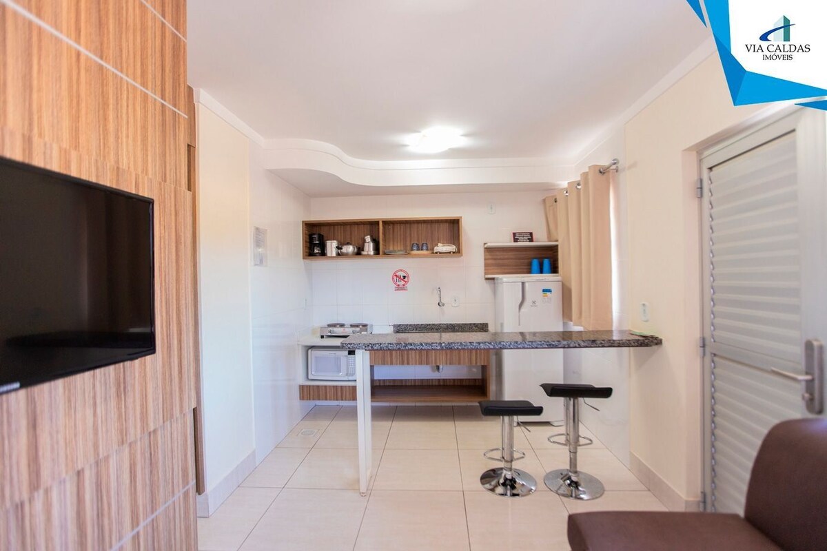 Cocina compacta con microondas, refrigerador pequeño y un mueble de madera. Hay un taburete y una silla en la zona de comedor.