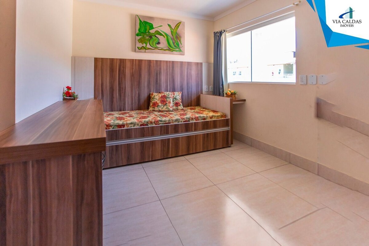 Un dormitorio con cabecera de madera, cama con ropa de cama floral, una mesita con un florero y una ventana con cortinas.
