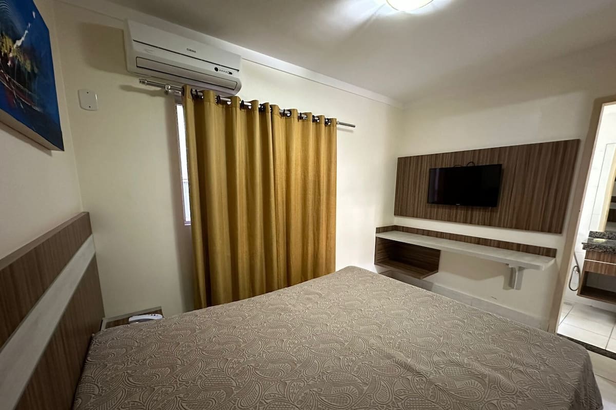 A hotel room with a bed, an air conditioning unit, a TV mounted on the wall, and a bathroom visible through an open door.