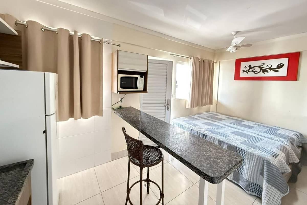 A compact room with a kitchenette, a bed with a checkered blanket, a dining table with a chair, and a ceiling fan.