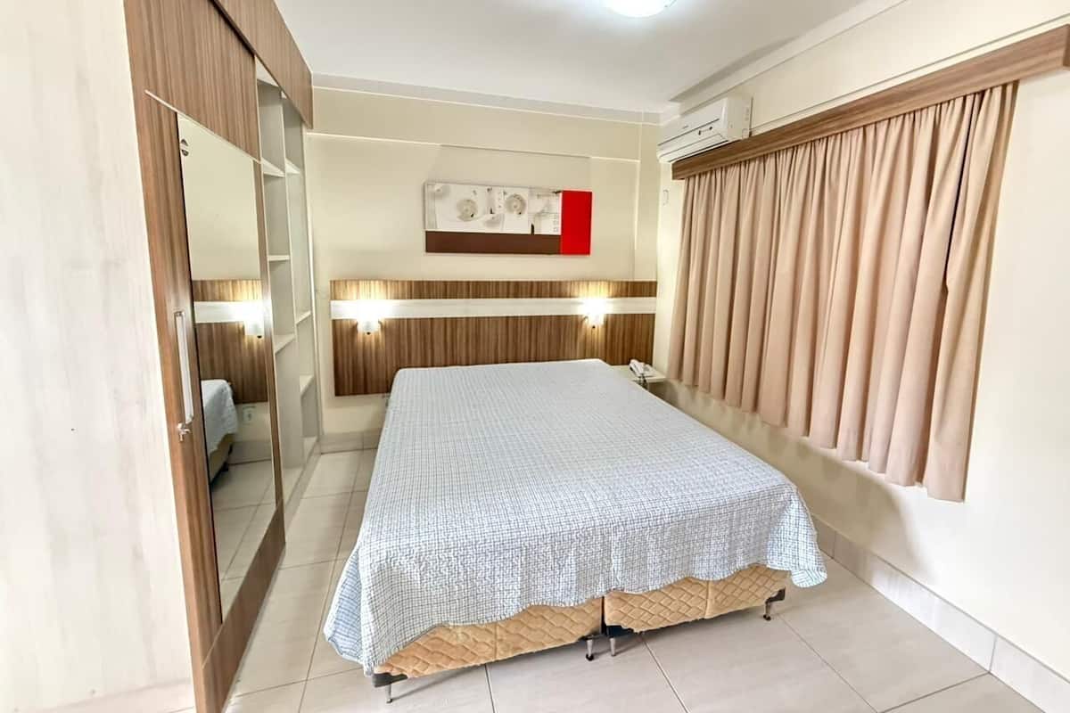 A bedroom with a bed, a wardrobe, a mirror, a wall-mounted air conditioner, and a window with curtains.