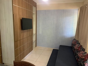 Quarto com televisão de tela plana fixada em parede de madeira, sofá estampado e piso de azulejos.