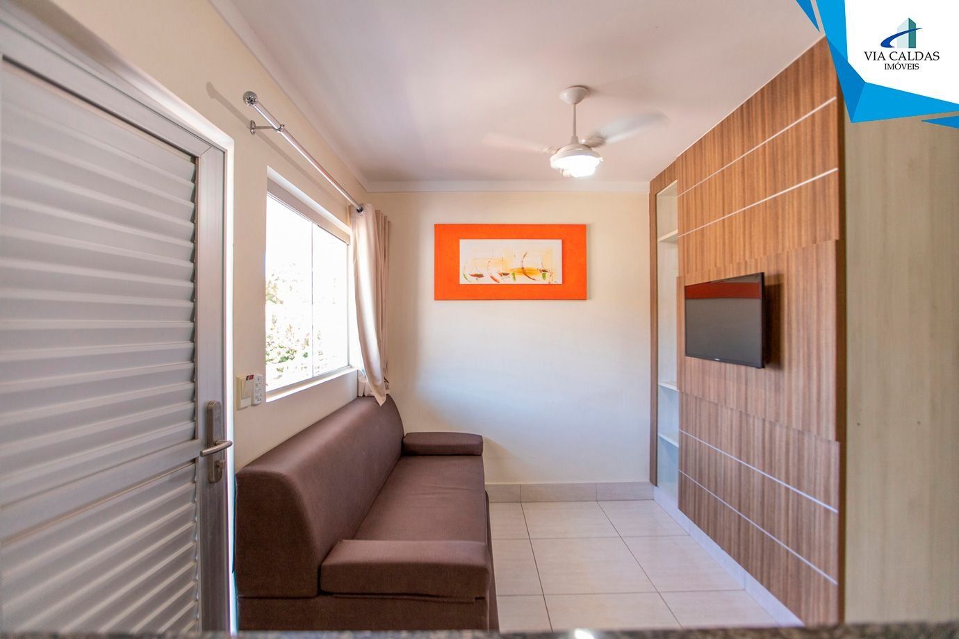 A room with a sliding door, a brown sofa, a ceiling fan, a framed picture on the wall, and a TV mounted on a wooden panel wall.