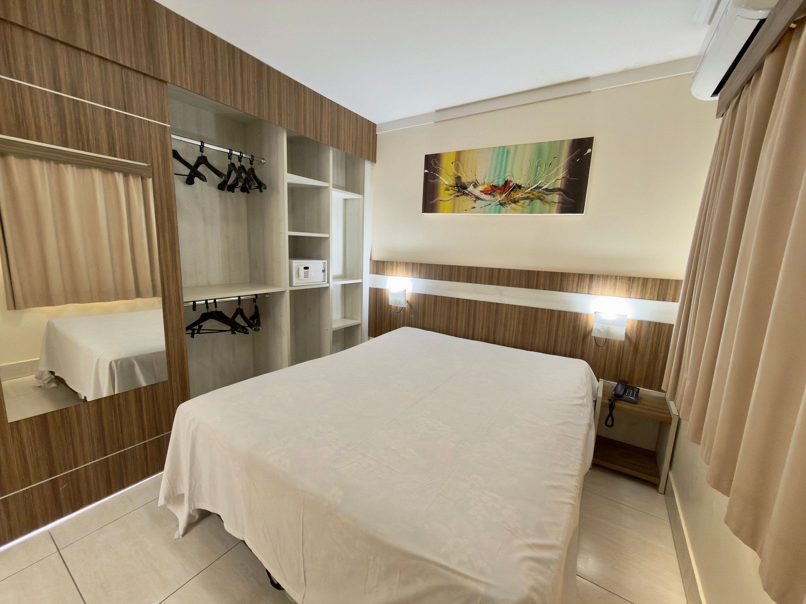 A hotel room with a bed, a wardrobe, a bedside table, a mirror, and a painting on the wall.