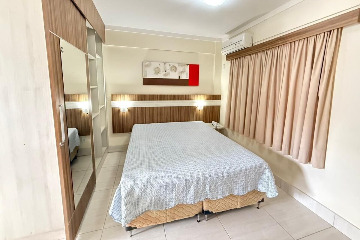 Un dormitorio con cama, armario, espejo, aire acondicionado instalado en la pared y ventana con cortinas.