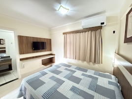Quarto de hotel com uma cama, televisão presa a um painel de madeira, janela com cortinas e ar condicionado.