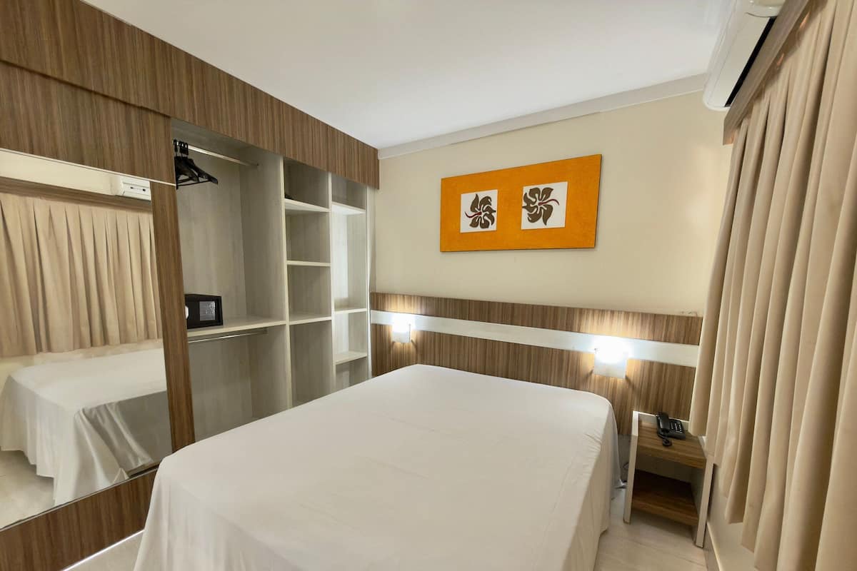 A hotel room with a bed, a wardrobe, a mirror, a bedside table, and a wall-mounted artwork.