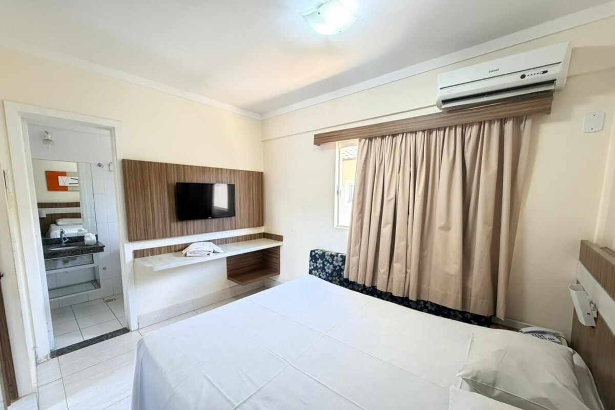 Habitación de hotel con cama, televisor, equipo de aire acondicionado, ventana con cortinas y un pequeño escritorio.