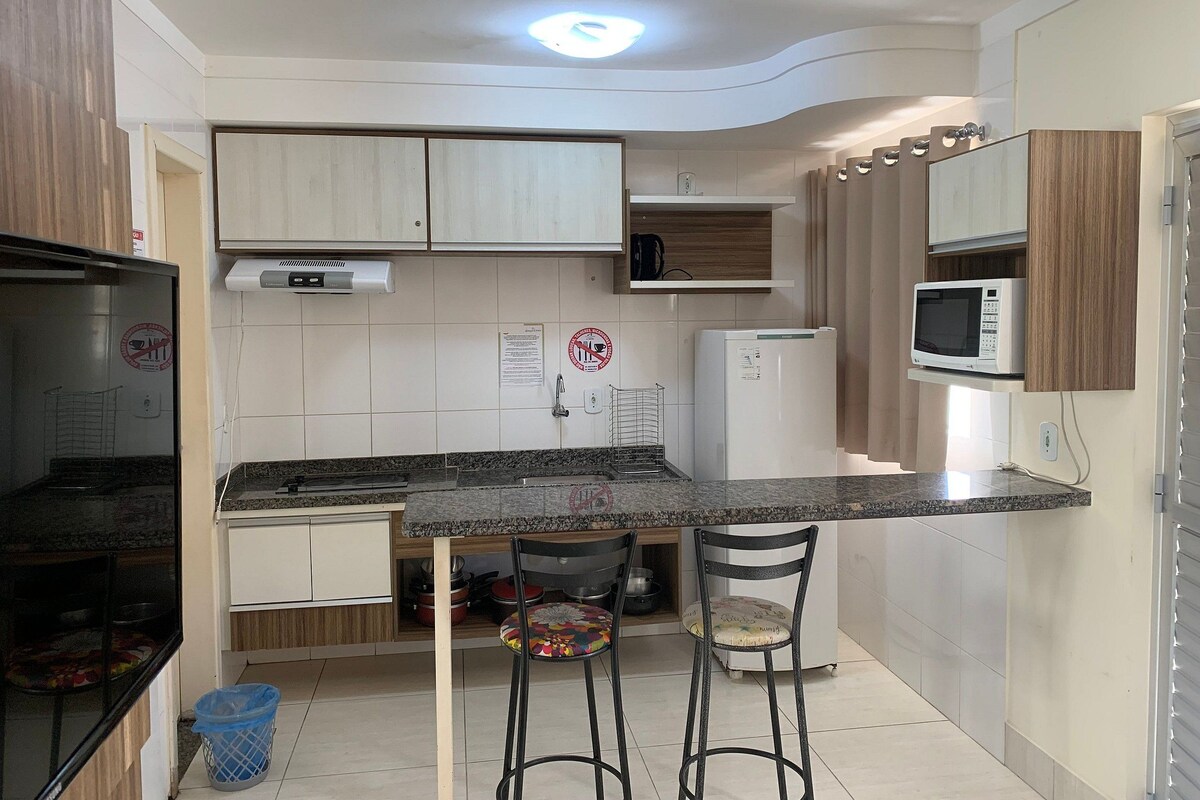 A compact kitchen with a built-in microwave, a refrigerator, and a sink. There are two bar stools and a small trash bin.