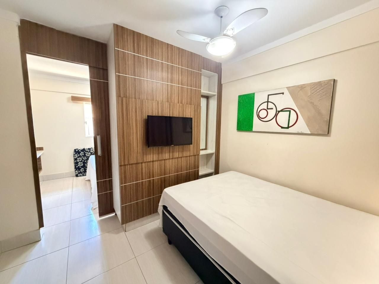 Quarto com cama, televisão, ventoinha no teto e obra de arte na parede.
