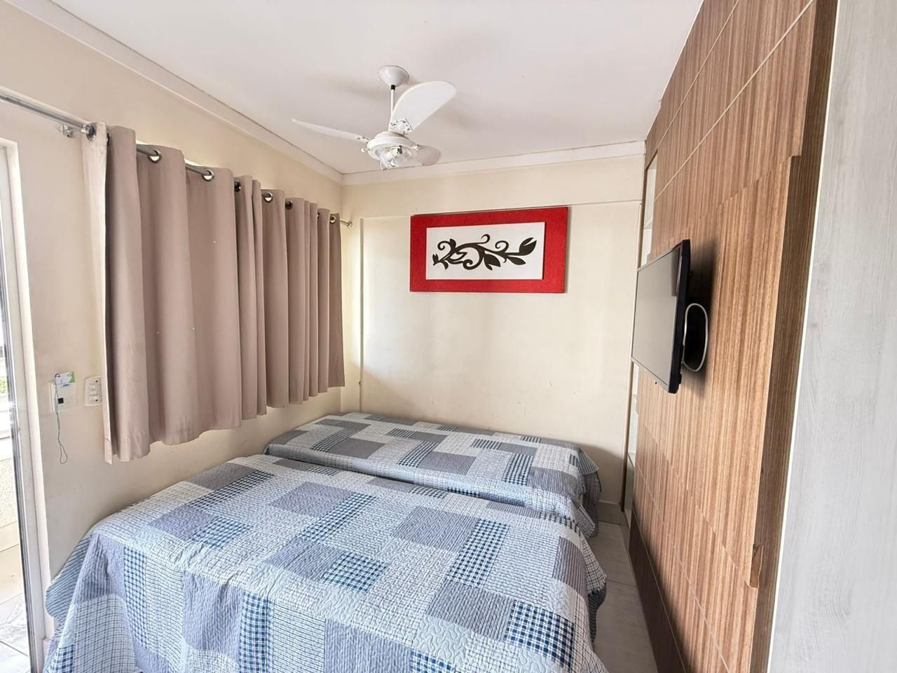 A bedroom with a bed, curtains, a ceiling fan, a wall-mounted TV, and a framed picture.
