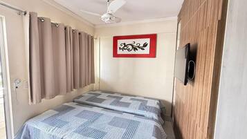 A bedroom with a bed, curtains, a ceiling fan, a wall-mounted TV, and a framed picture.