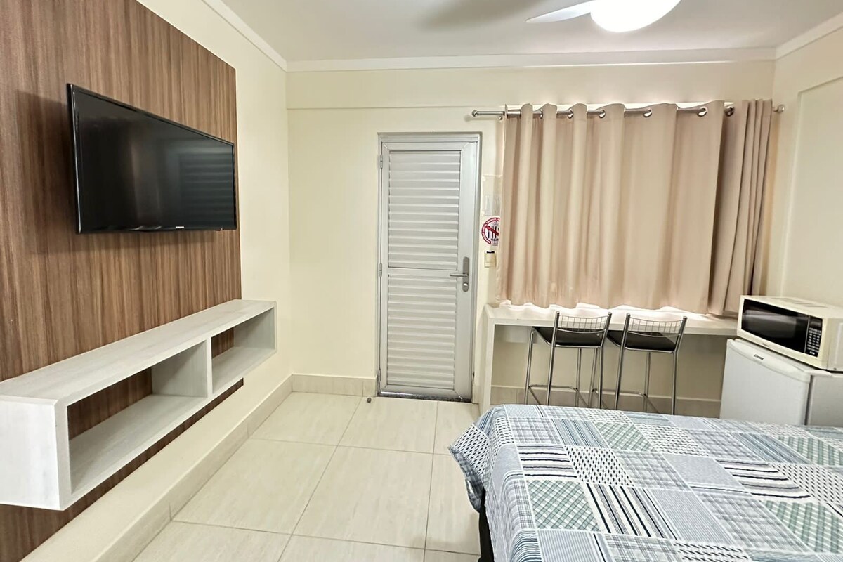 Un dormitorio con cama, televisor, escritorio con sillas, microondas y ventilador de techo.