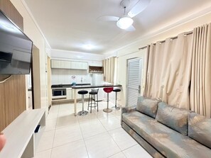 A modern living room with a sofa, a TV, a ceiling fan, and a kitchen area with a bar stool and a microwave. - Lacqua DiRoma II Via Caldas (Caldas Novas)