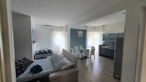 A modern living room with a sofa, a dining table, and chairs. There is a kitchen area with cabinets and a refrigerator.
