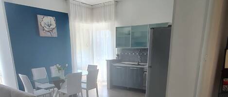 Una sala de estar moderna con sofá, mesa de comedor y sillas. Se observa en el fondo una zona de cocina con armarios y electrodomésticos.