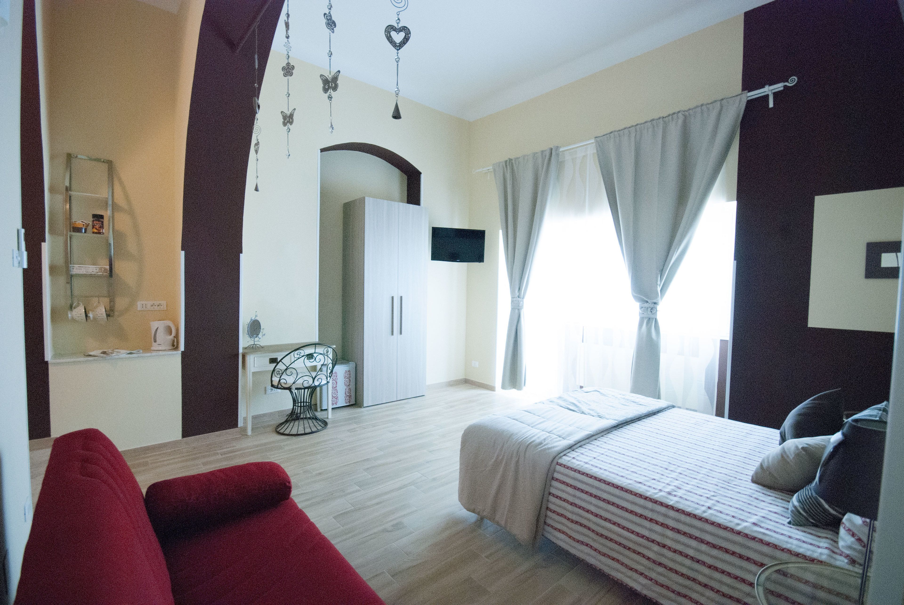 A hotel room with a bed, a red chair, a desk with a chair, a TV, and a wardrobe.