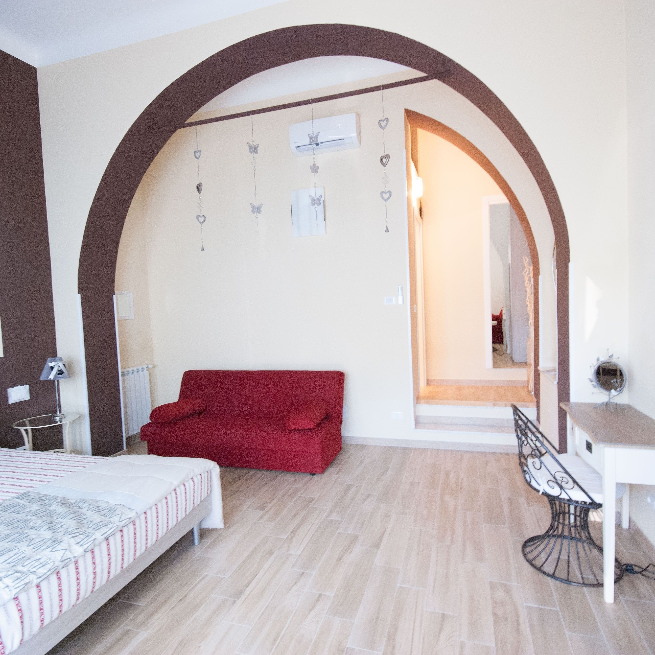 A bedroom with a red sofa, a bed with a patterned bedspread, a desk with a chair, and a large arched doorway leading to another room.