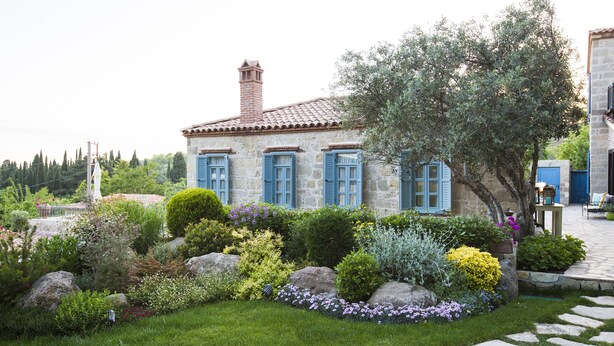 A stone house with blue shutters, surrounded by a well-maintained garden with various plants and a stone pathway. - Ida Blue, Adatepe - Adults Only (Ayvacik)