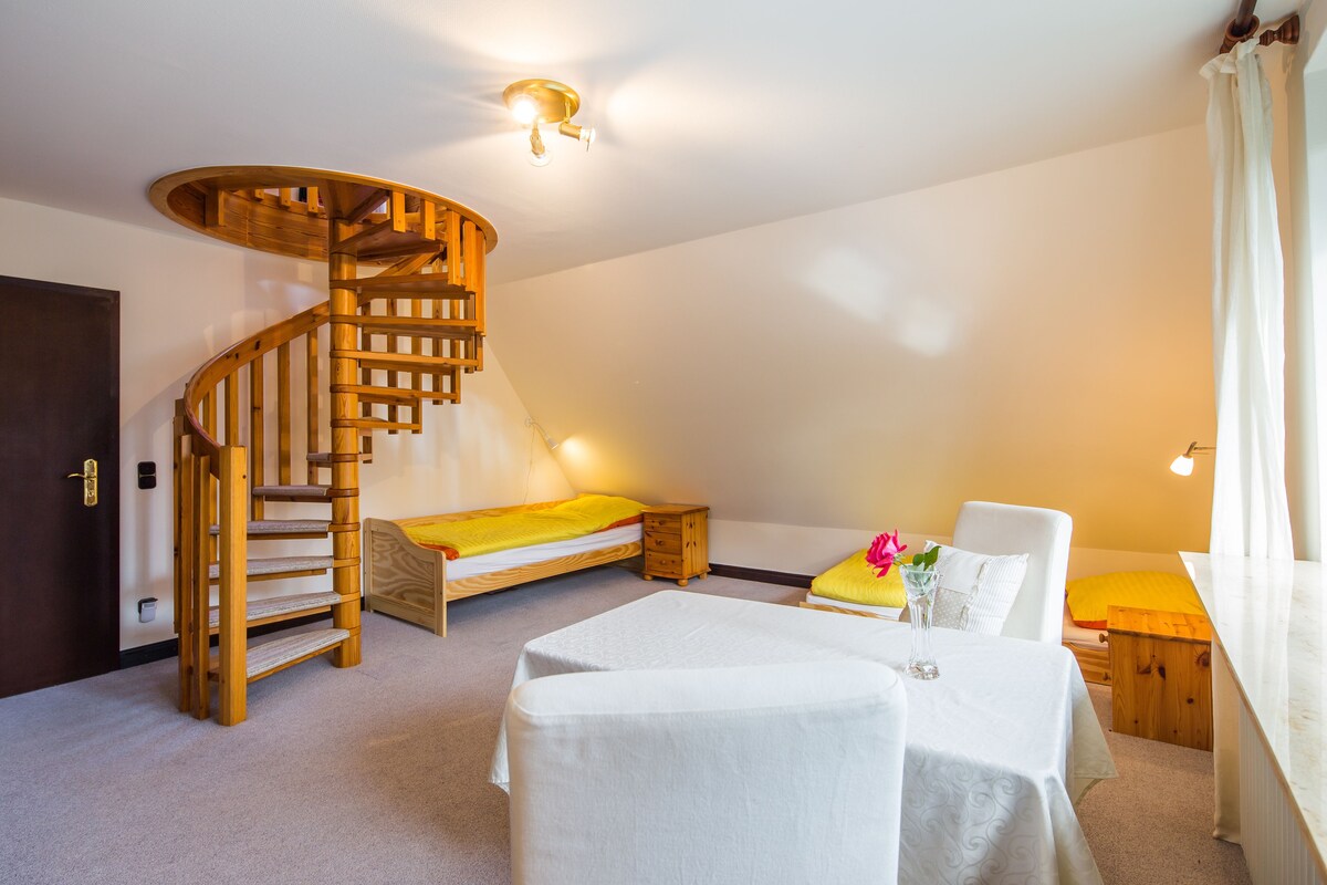 Een slaapkamer met een houten spiraaltrap, een bed, een klein tafeltje en een raam met gordijnen.