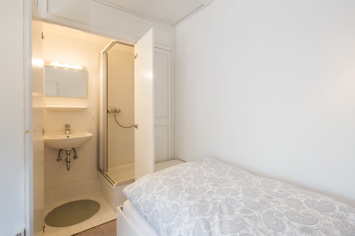 Een slaapkamer met een bed, een badkamer met wastafel en douche, en een deur die leidt naar een andere kamer.