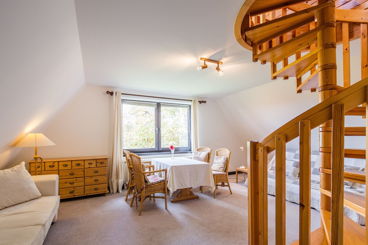 Een kamer met een houten trap, een eettafel met stoelen, een bank en een kledingkast.