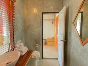 Un baño con tocador de madera, lavamanos blanco y retrete. Hay un perchero con toallas dobladas y una pequeña cesta. Las paredes tienen textura y una puerta da a otra habitación.