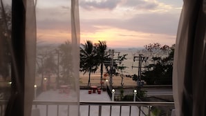 Un balcon offrant une vue sur le coucher de soleil sur la mer, avec des palmiers et un espace salon extérieur.