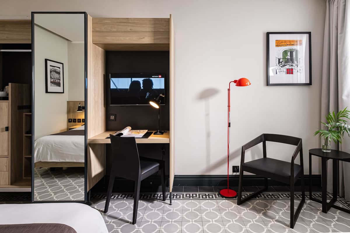Une chambre d’hôtel compacte comprenant un lit, un bureau avec un ordinateur, une chaise, une lampe de sol rouge et une petite table avec une plante.