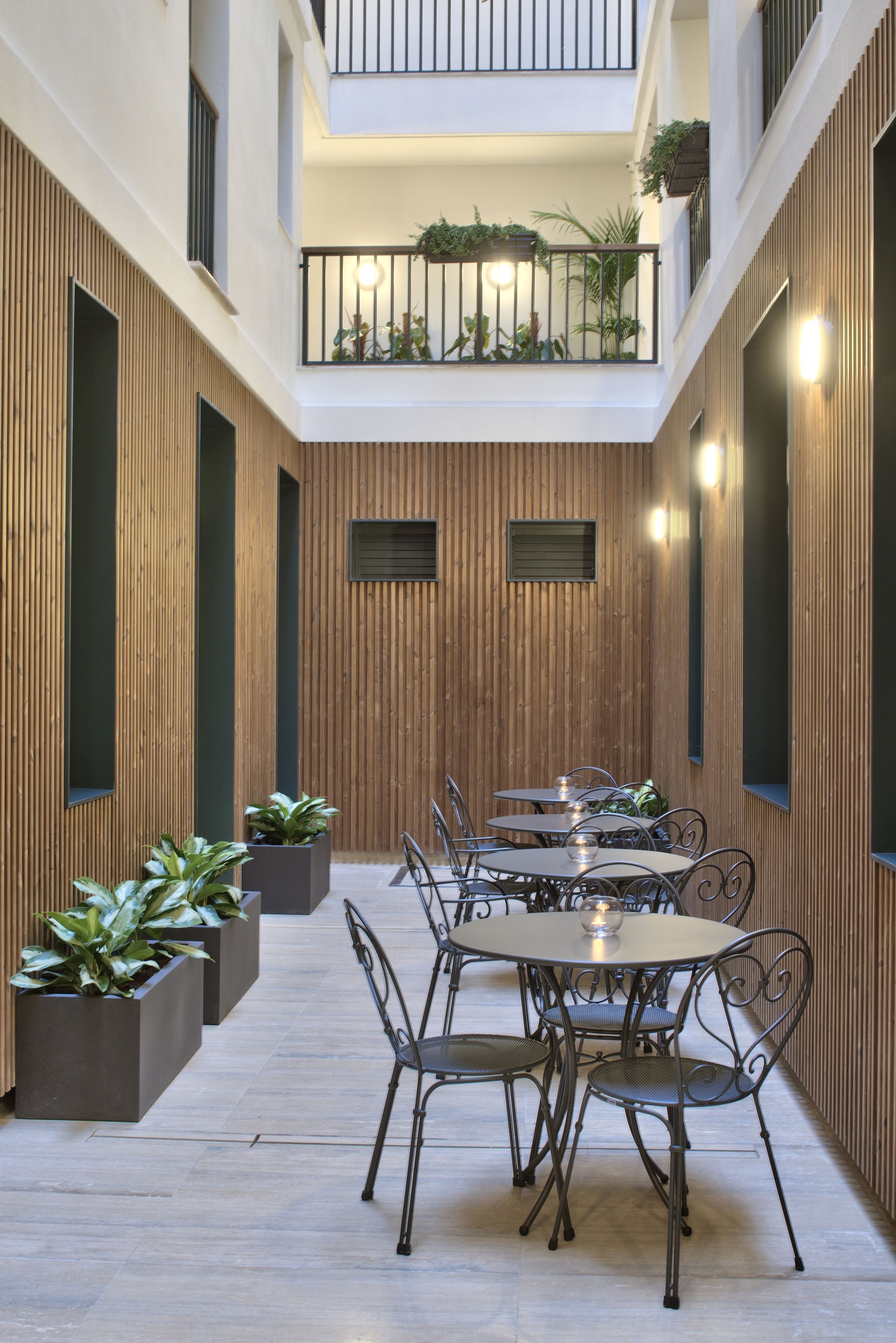 A modern indoor courtyard with a wooden wall, a balcony with plants, and a dining area with metal chairs and tables.