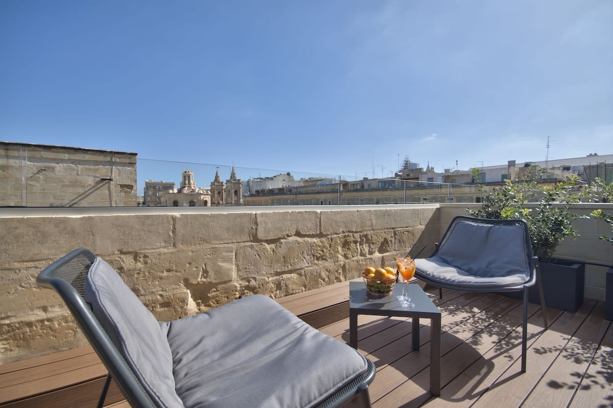 Une terrasse sur le toit avec deux chaises longues, une petite table avec des fruits et une boisson, et une vue sur des bâtiments historiques en arrière-plan.