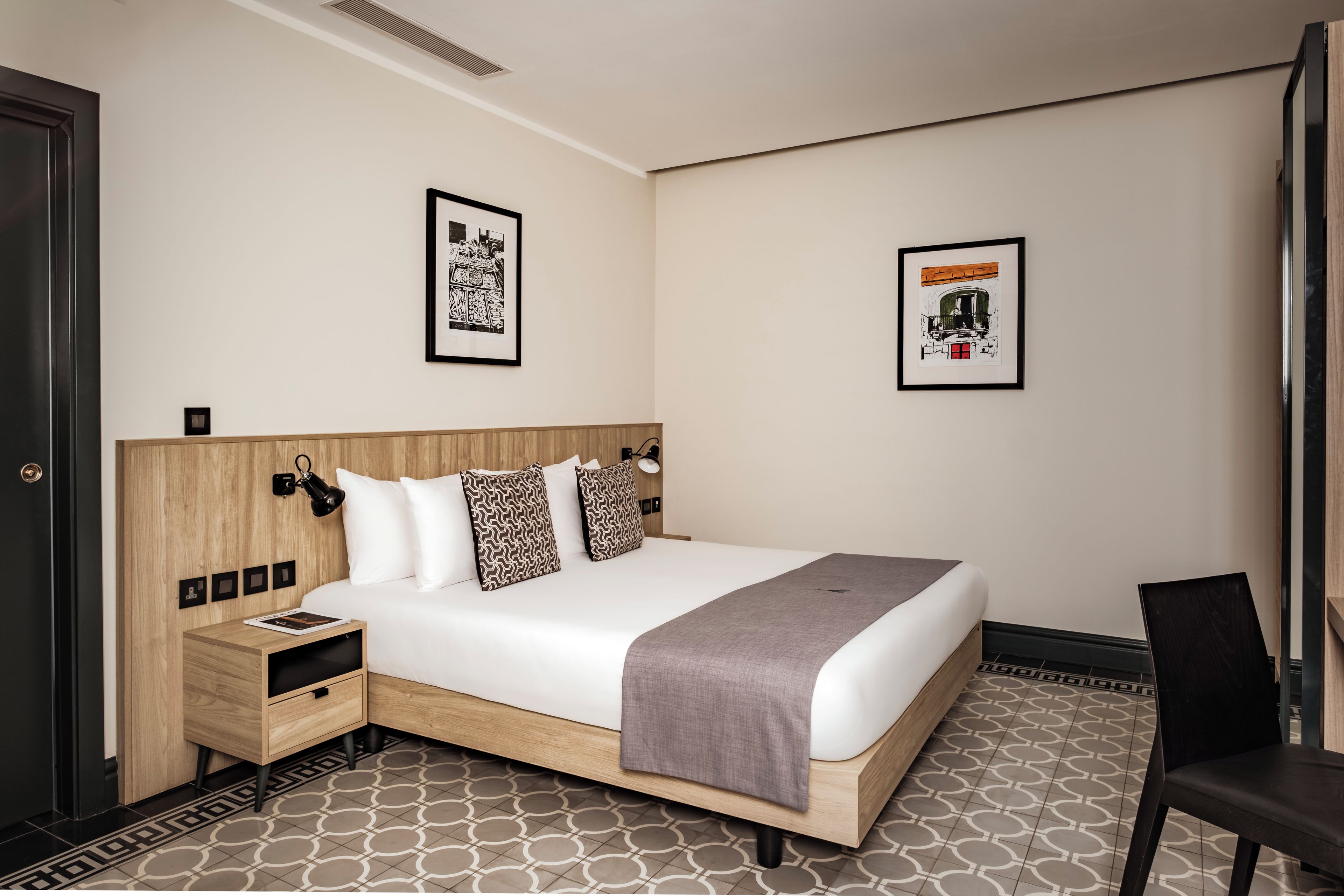 A hotel room with a bed, bedside tables, a chair, and two framed pictures on the wall.